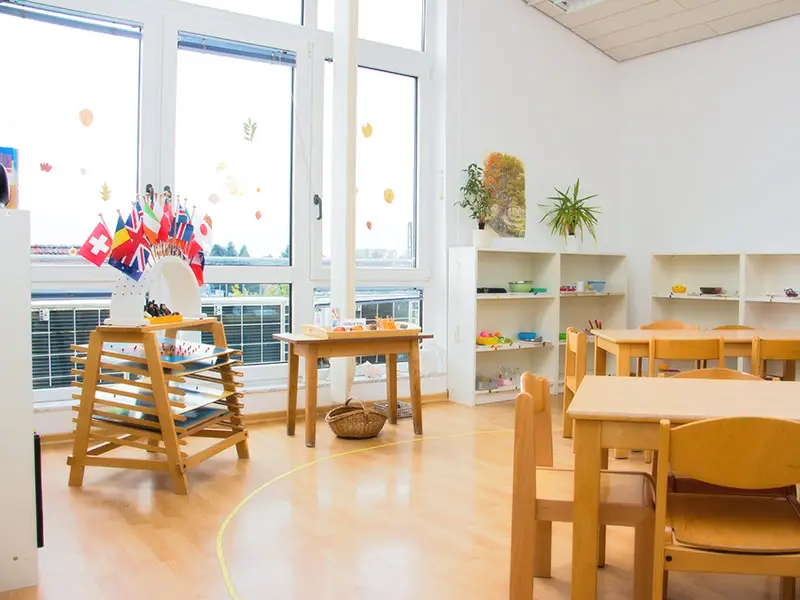 Heller Gruppenraum mit Montessori-Regalen, Kindertischen und internationalen Fahnen